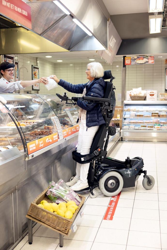 A white female in a Balder Finesse F390. She is in the standing position in a grocery store. She is reaching across the counter to a store assistant.