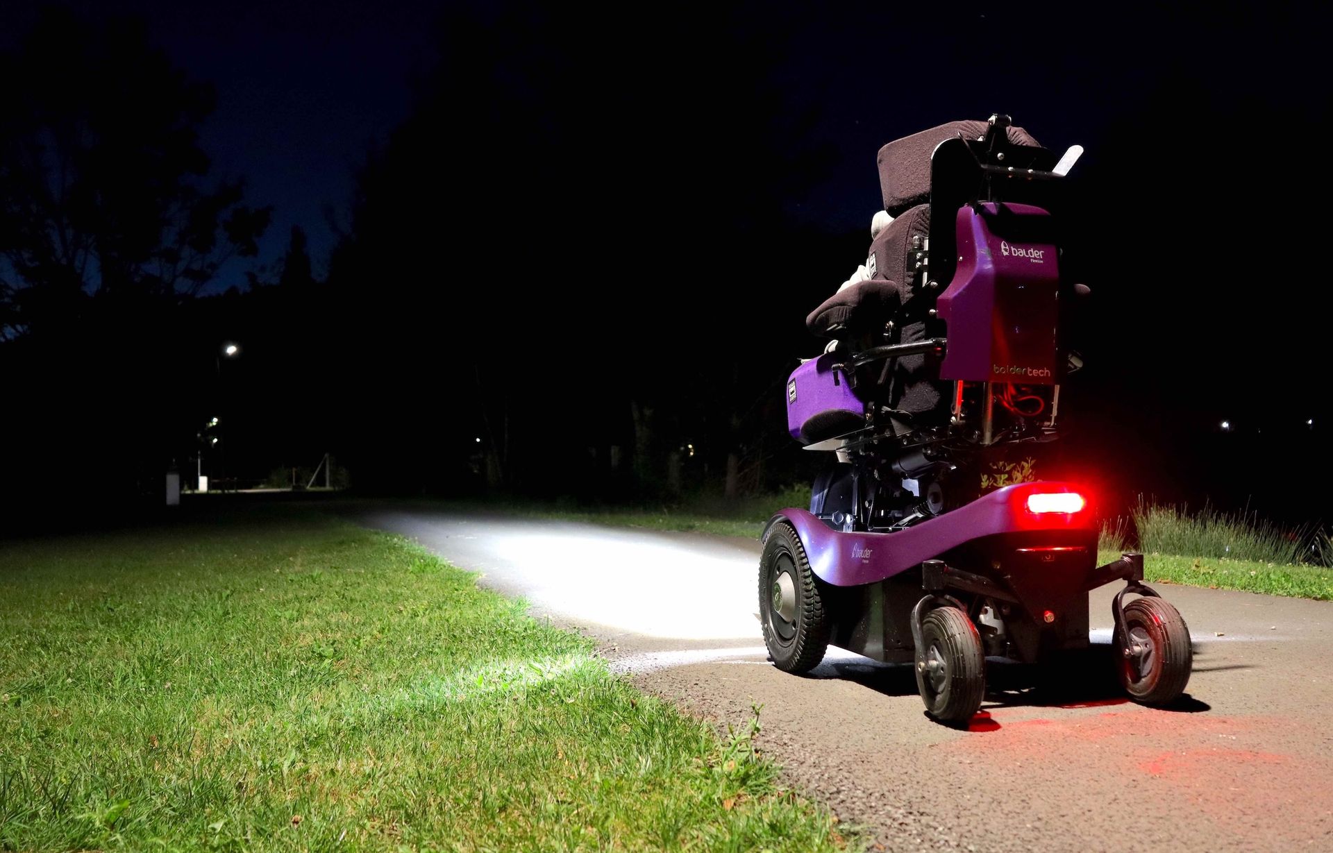A purple Balder Finesse powerchair driving along a path at night. A red LED light is visible on the back of the wheelchair, while headlights illuminate the path ahead.